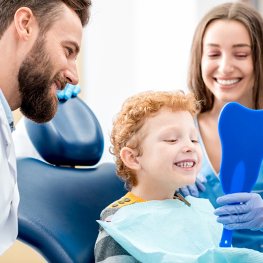 a young child looking at his teeth in a tooth-shaped mirror held by a dental assistant